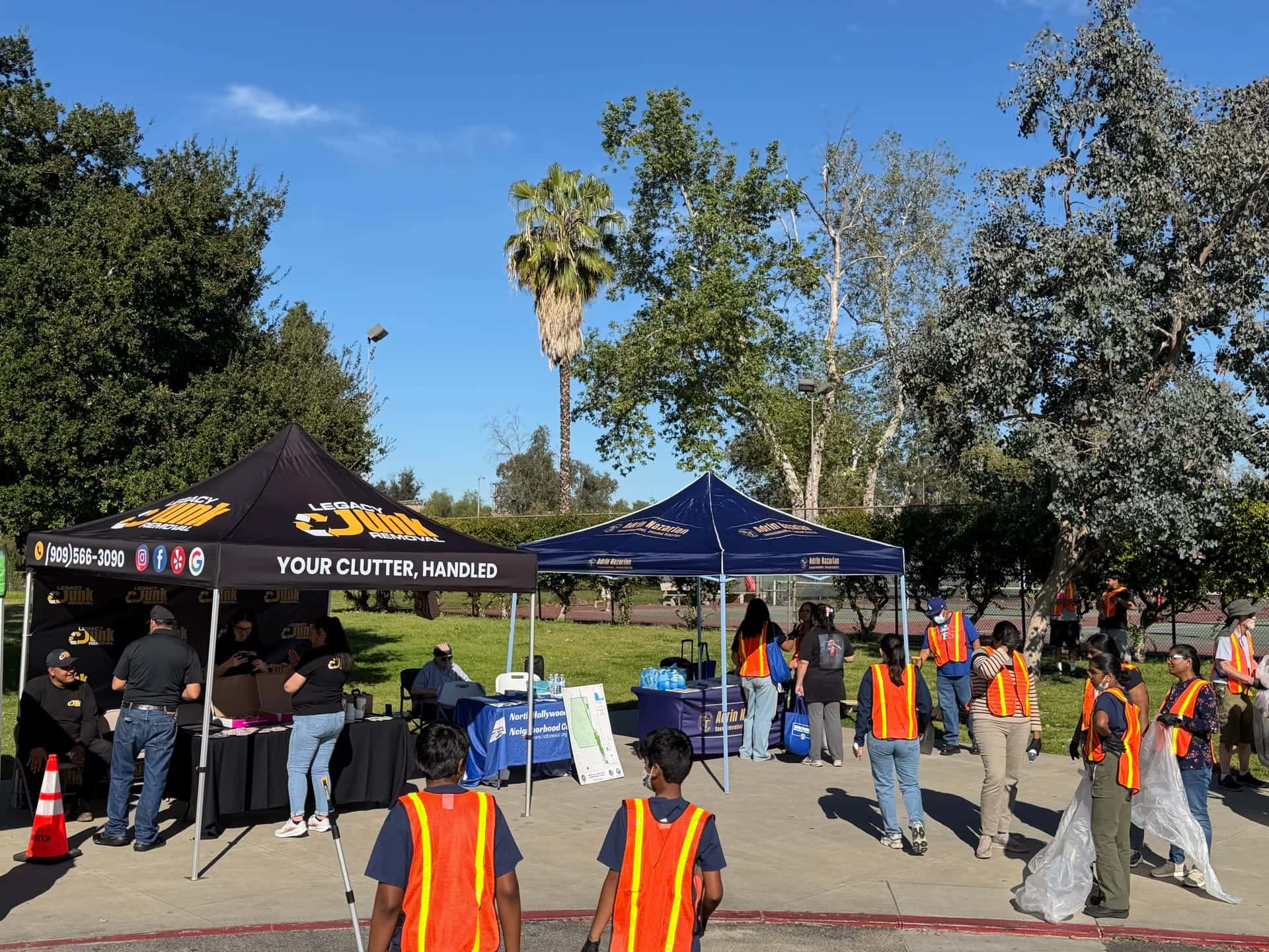 The Legacy canopy and a partner booth in a park during a community clean-up, with volunteers in orange safety vests
