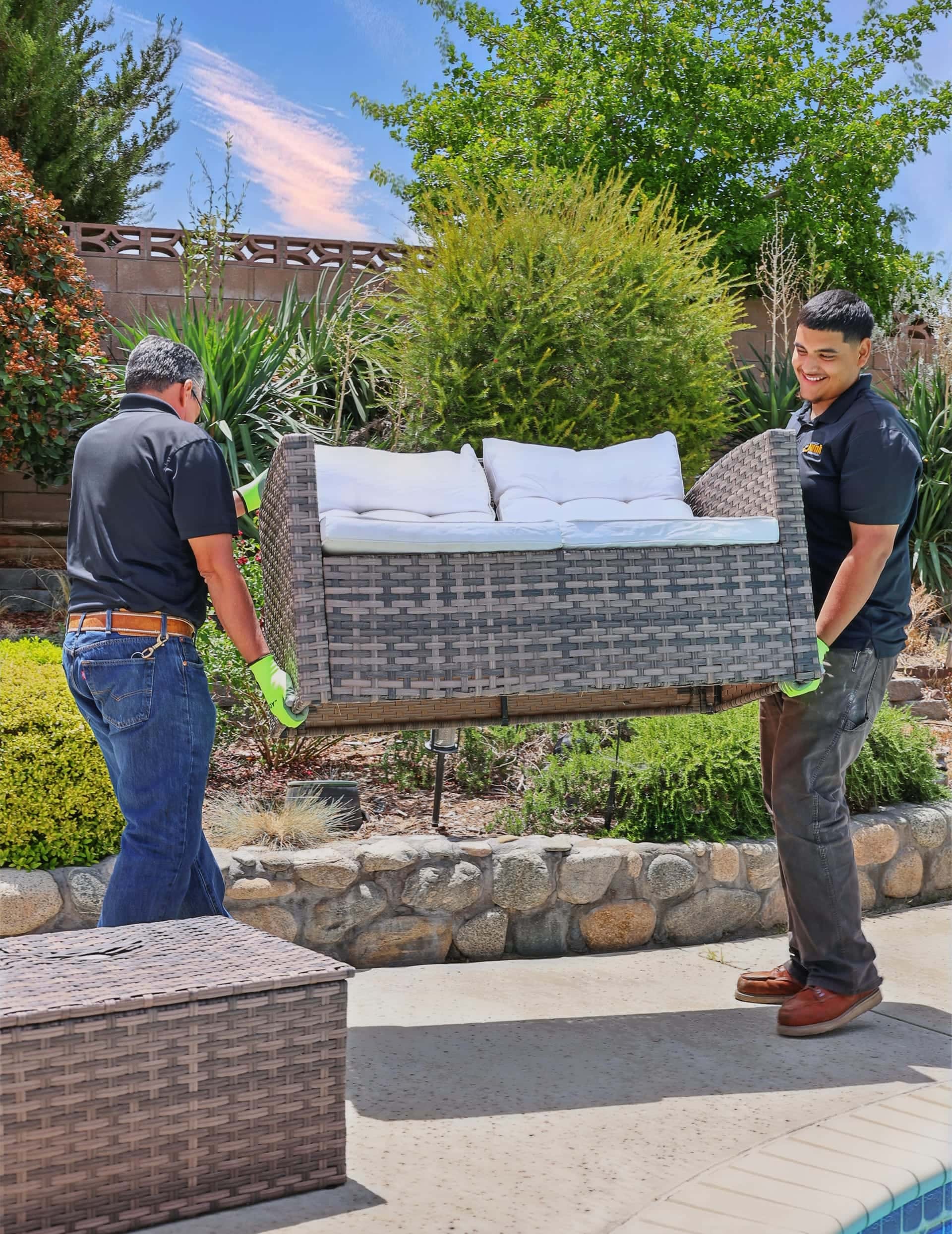 Two Legacy crew members carrying a wicker patio sofa past a pool