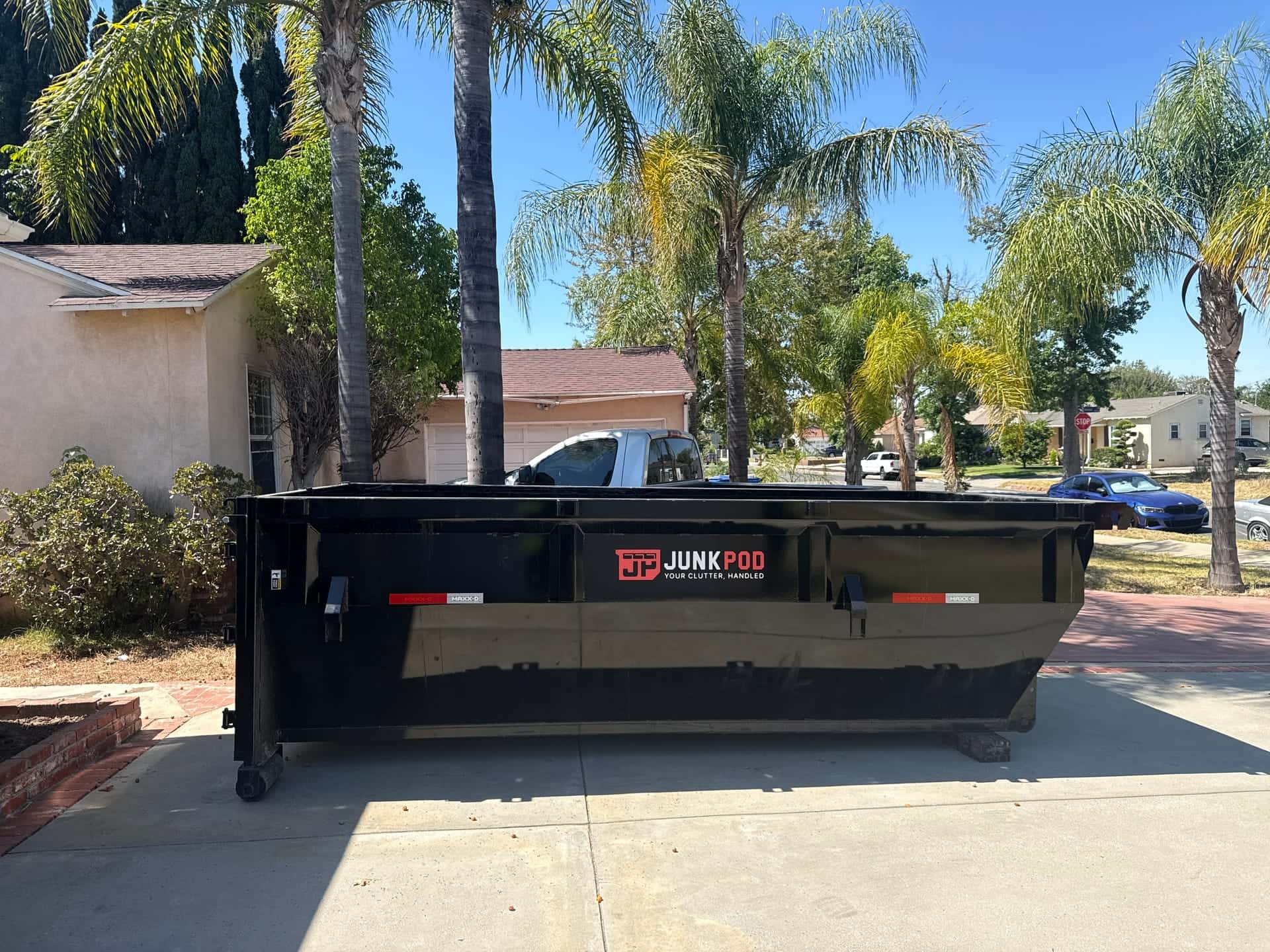 A black Legacy dumpster in a residential driveway on a palm-lined street